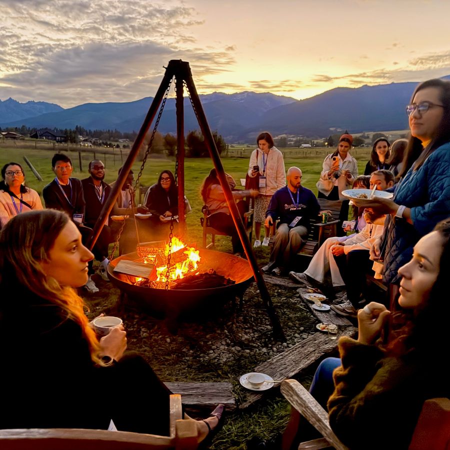 a large group of young professionals sitting outdoors in a circle around a western style firepit in a metal caldron suspended from a wooden tripod, in front of the Northern Rocky Mountains with a glowing golden sunset
