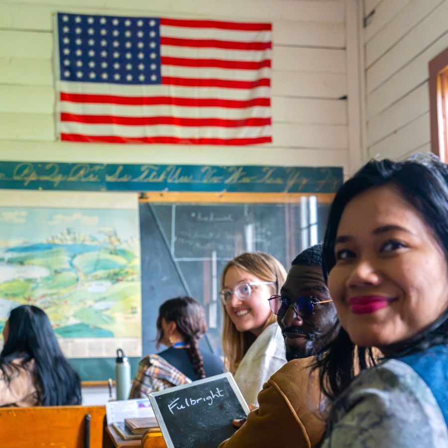 Several young professionals sitting in a restored historic one-room schoolroom with an American Flag on the wall and an antique style map and chalkboard and painted wooden clapboard paneling
