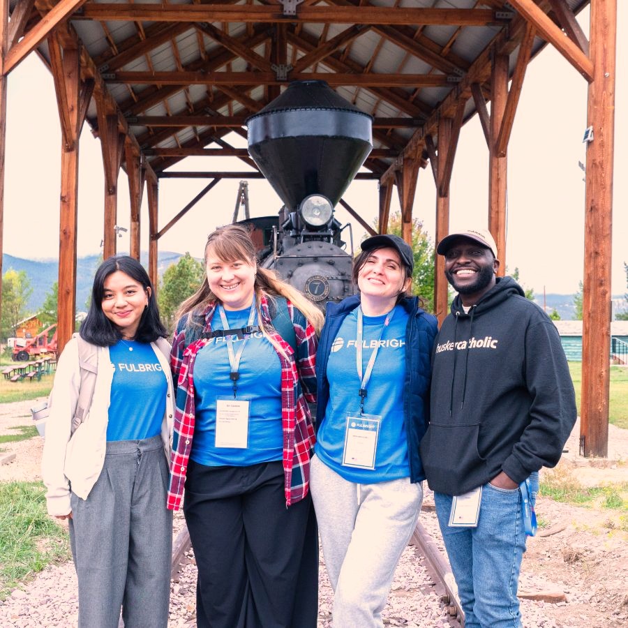 Four young adults wearing blue t-shirts that say Fulbright, standing in front of an antique train in front of a mountain setting