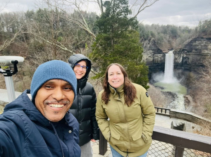 Man standing with two women in front of evergreen trees and rocky gorge with a waterfall, on a viewing terrace with a viewing telescope