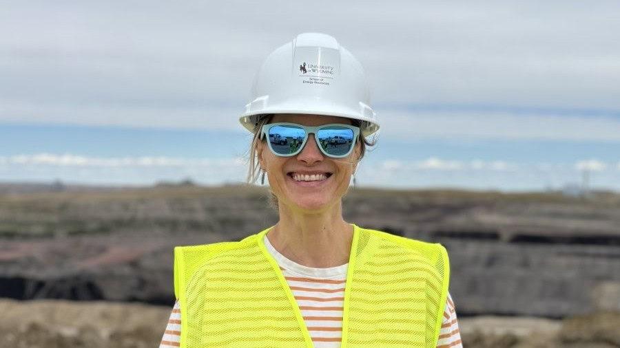 Woman wearing a white hard-hat safety helmet, sunglasses, and a high visibility neon yellow vest in an outdoor location with rocks and water