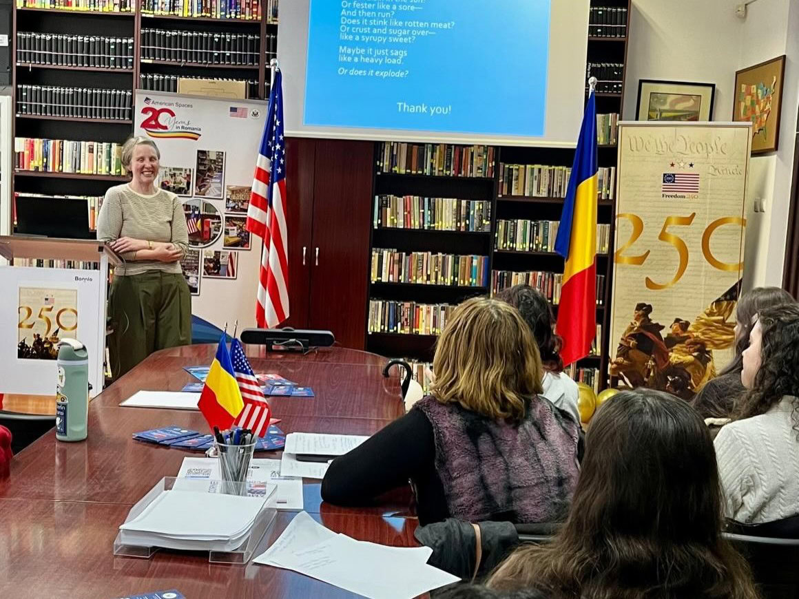 Woman standing at front or a room with American flag beside her and bookshelves behind her speaking to students in a library style room with a large conference table