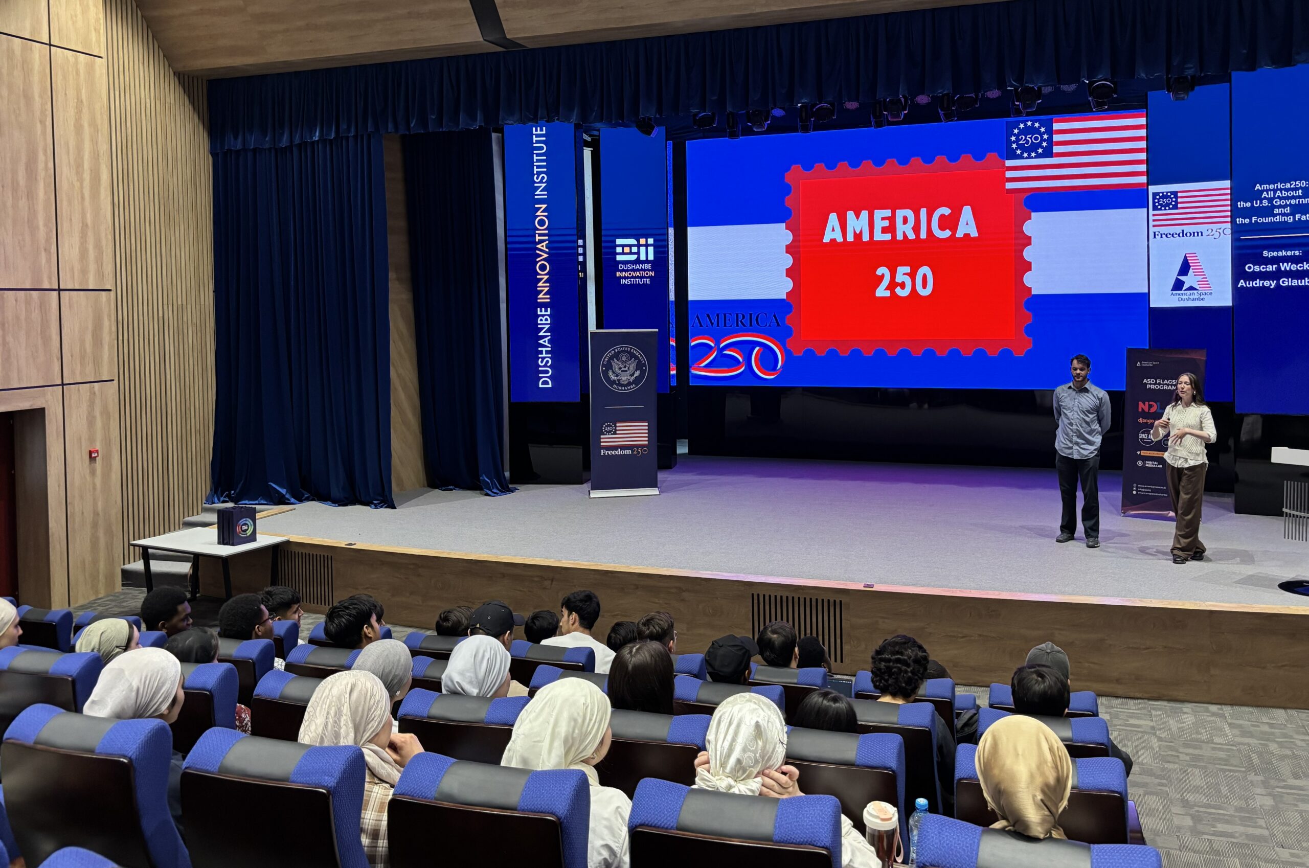 Two American students standing on a stage facing students sitting in theater style seats, with projection of American flag images and America 250 in red, white and blue on screen