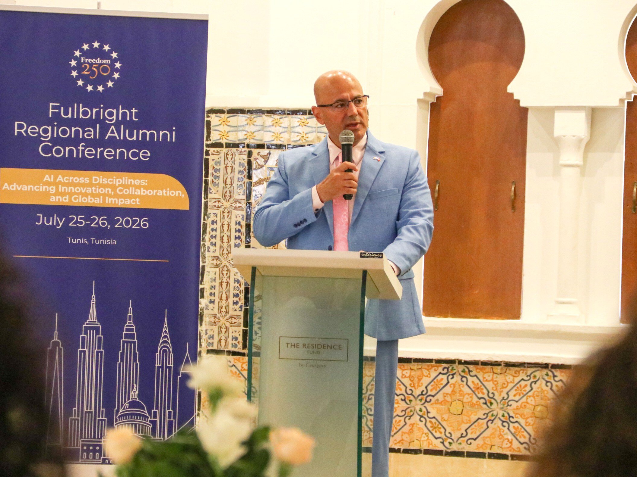Man in suit at podium holding microphone standing in fronot of a banner and a tiled wall with Tunisian tile patters and architecture