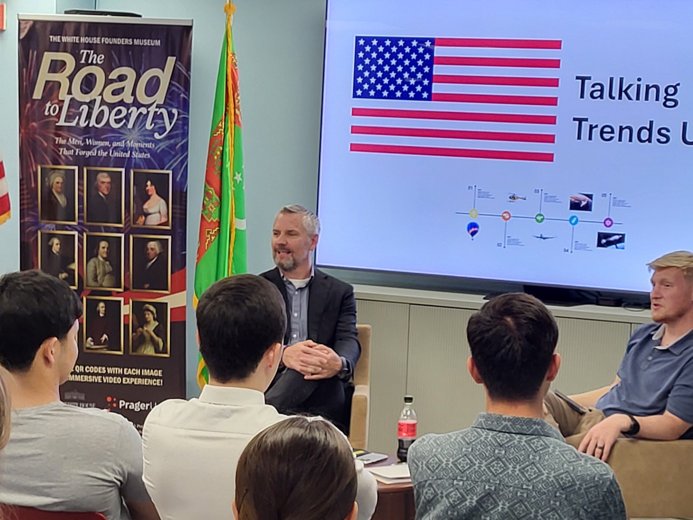 Man sitting at front of room speaking to students with banner and screen behind him with American flag and American historical photos